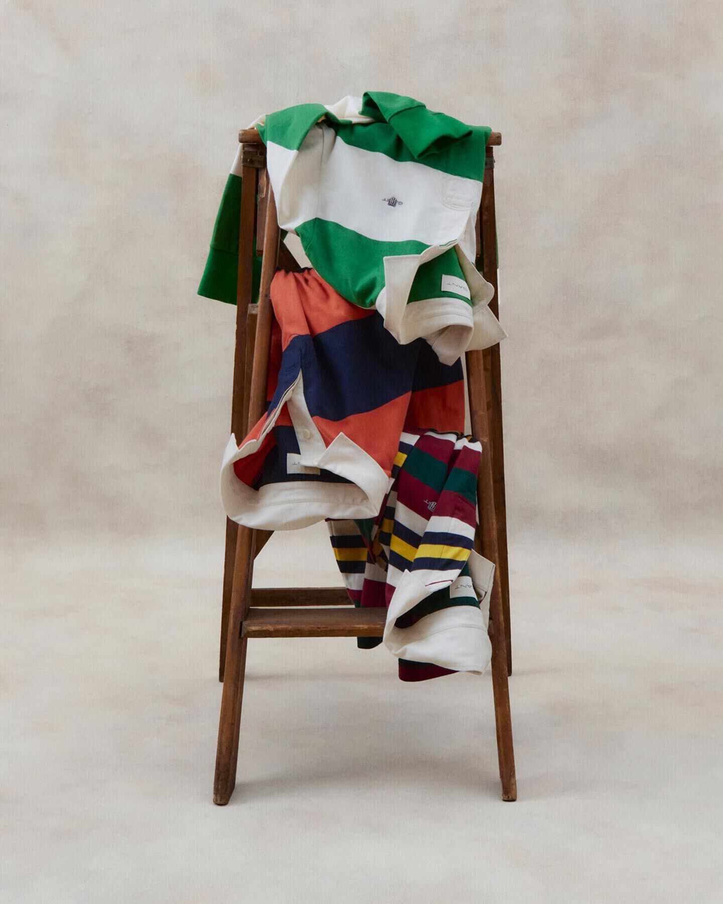 Three bold Gant Ruggers draped on a wooden stool—laid-back layers in a clean, minimal setting